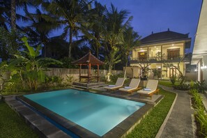 A swimming pool with lounge chairs and a gazebo, surrounded by lush greenery and palm trees, with a two-story house in the background. - Pondok Ayu Shanti (Ubud)