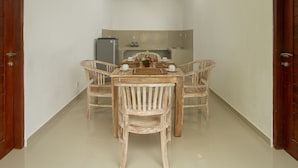 A dining area with a wooden table and four wicker chairs, a small kitchen area in the background, and a tiled floor. - Pondok Ayu Shanti (Ubud)