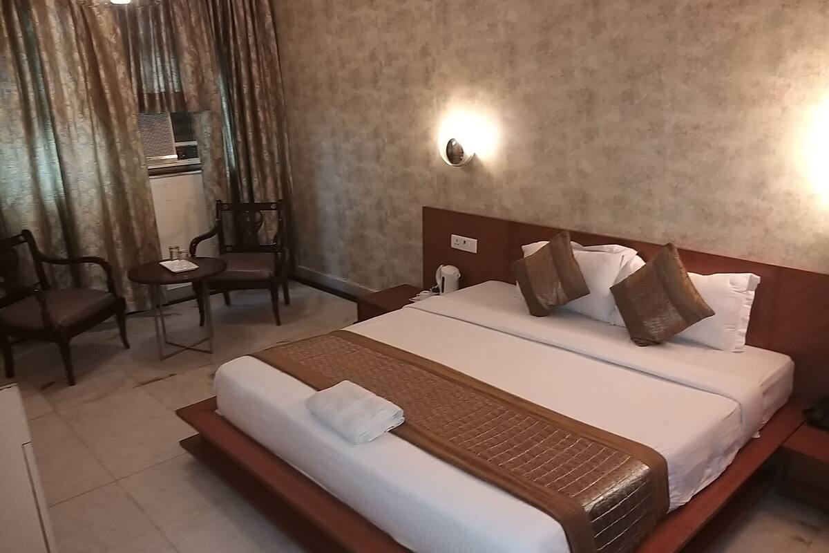 Habitación de hotel con una cama grande, una mesita de noche, una silla y una pequeña mesa con una tetera.