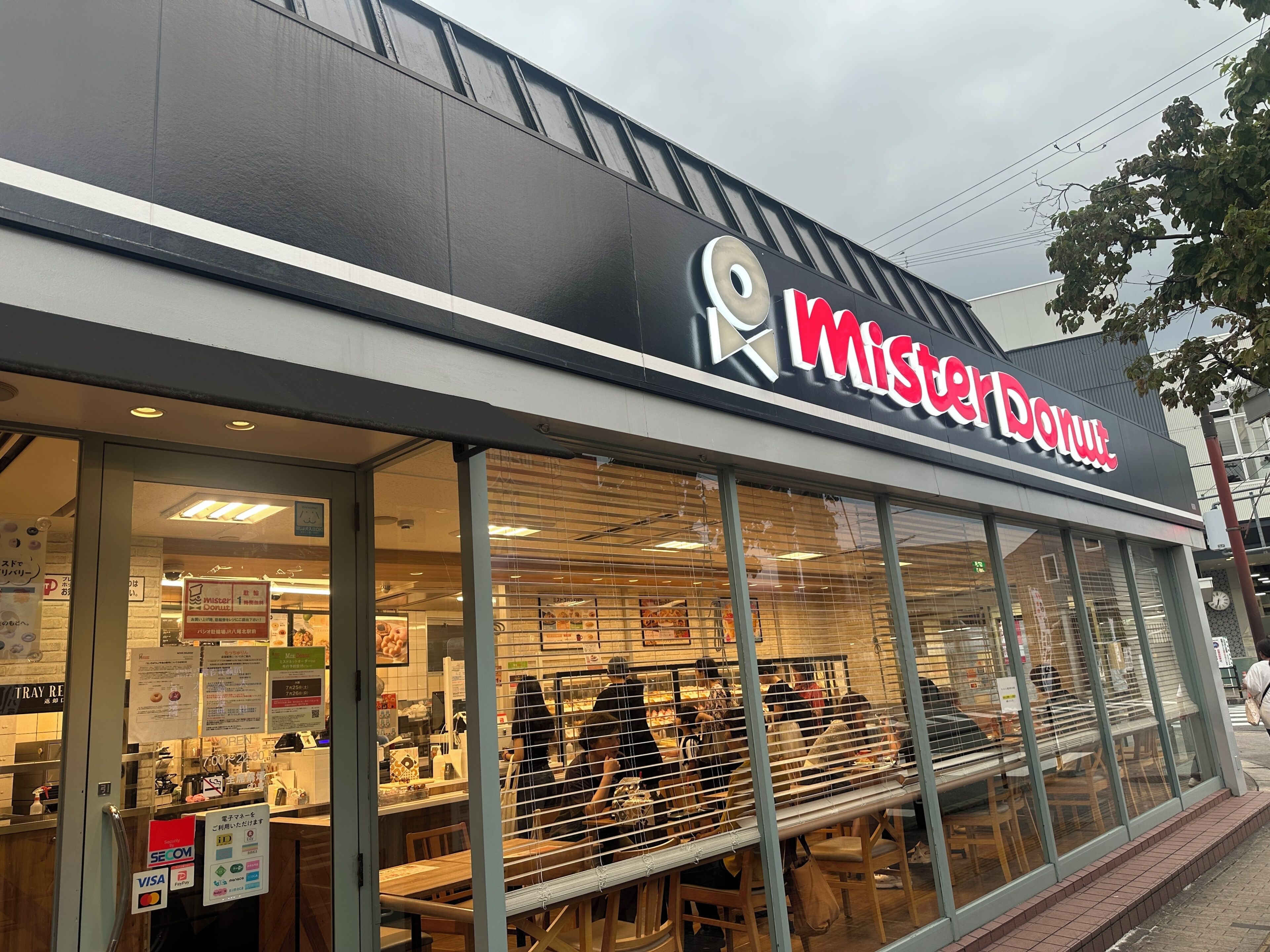 Una tienda Mister Donut con un amplio escaparate de vidrio, carteles de menú visibles y una vista clara del área de asientos interior.