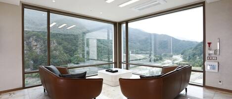 Una habitación moderna con ventanas amplias, dos sofás de cuero marrón, una mesa de centro blanca y vistas a un paisaje montañoso.
