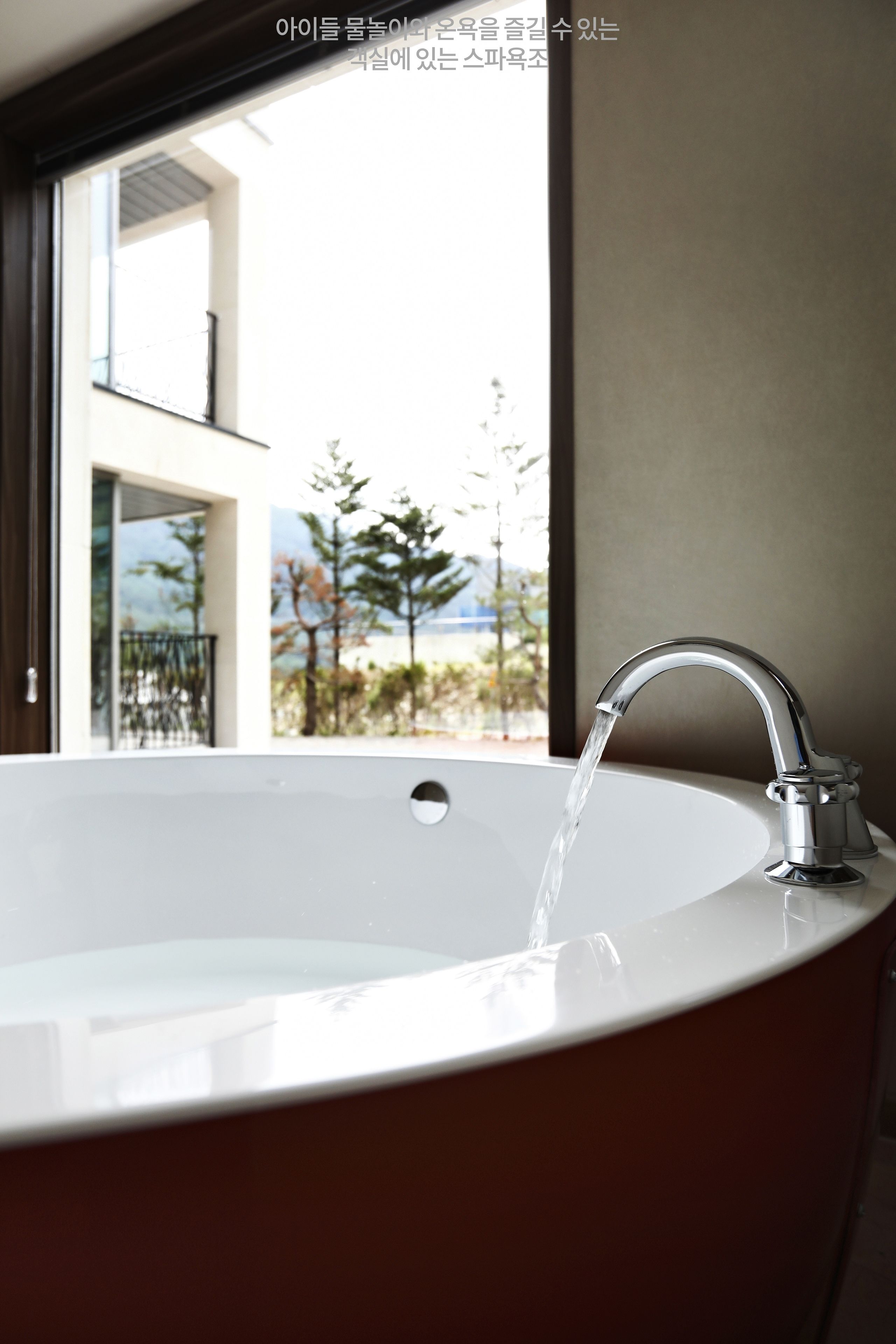 Un baño moderno con una bañera grande, una ventana con vistas a árboles y un edificio, y un grifo con agua corriendo.