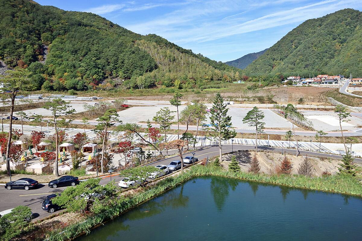A parking lot with cars, a small pond, and a road curving through a hilly area with trees.