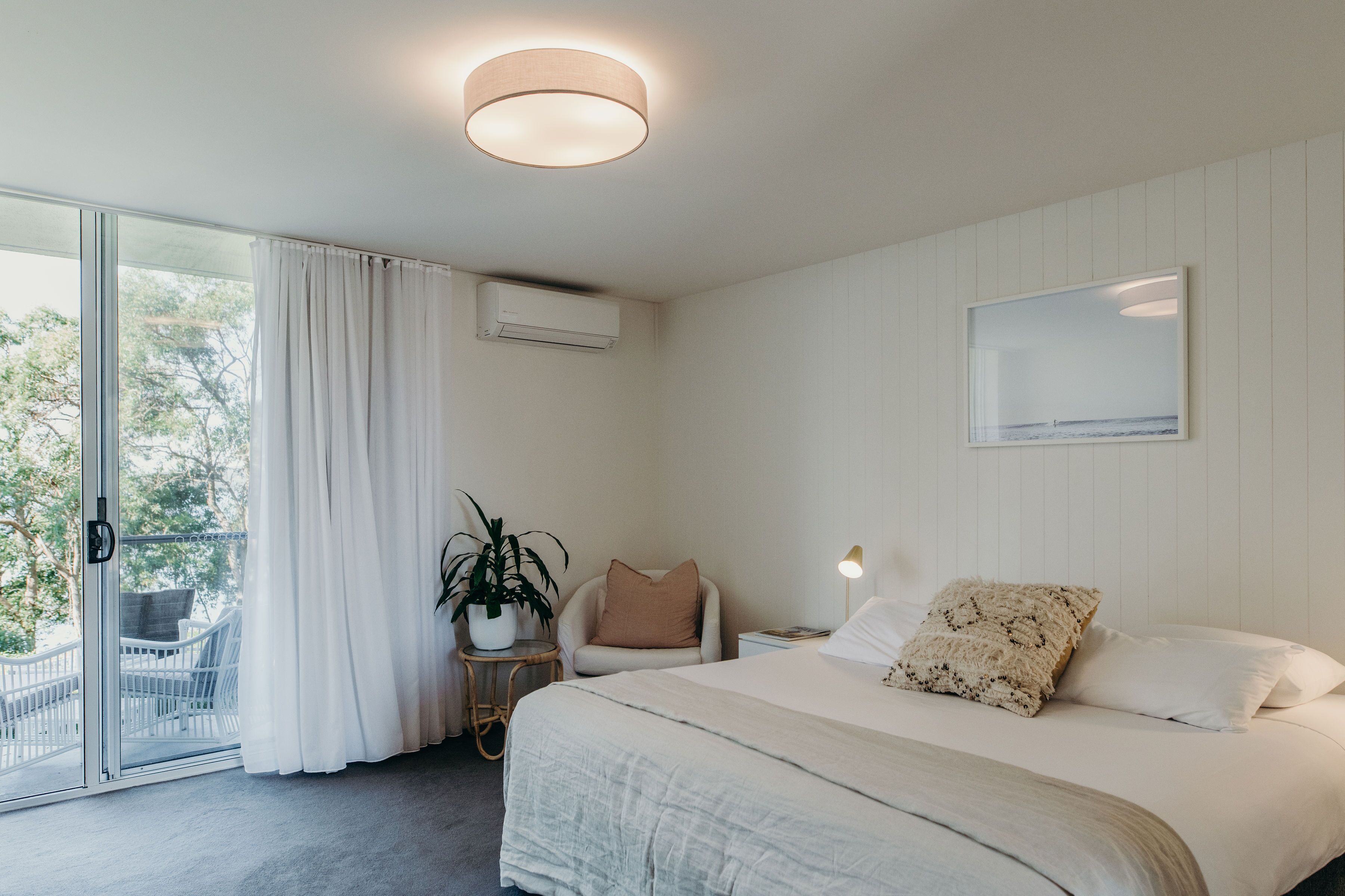 Ein Schlafzimmer mit Bett, Nachttisch, Lampe, Stuhl, einer Topfpflanze und einer Schiebeglastür, die zu einem Außenbereich führt.