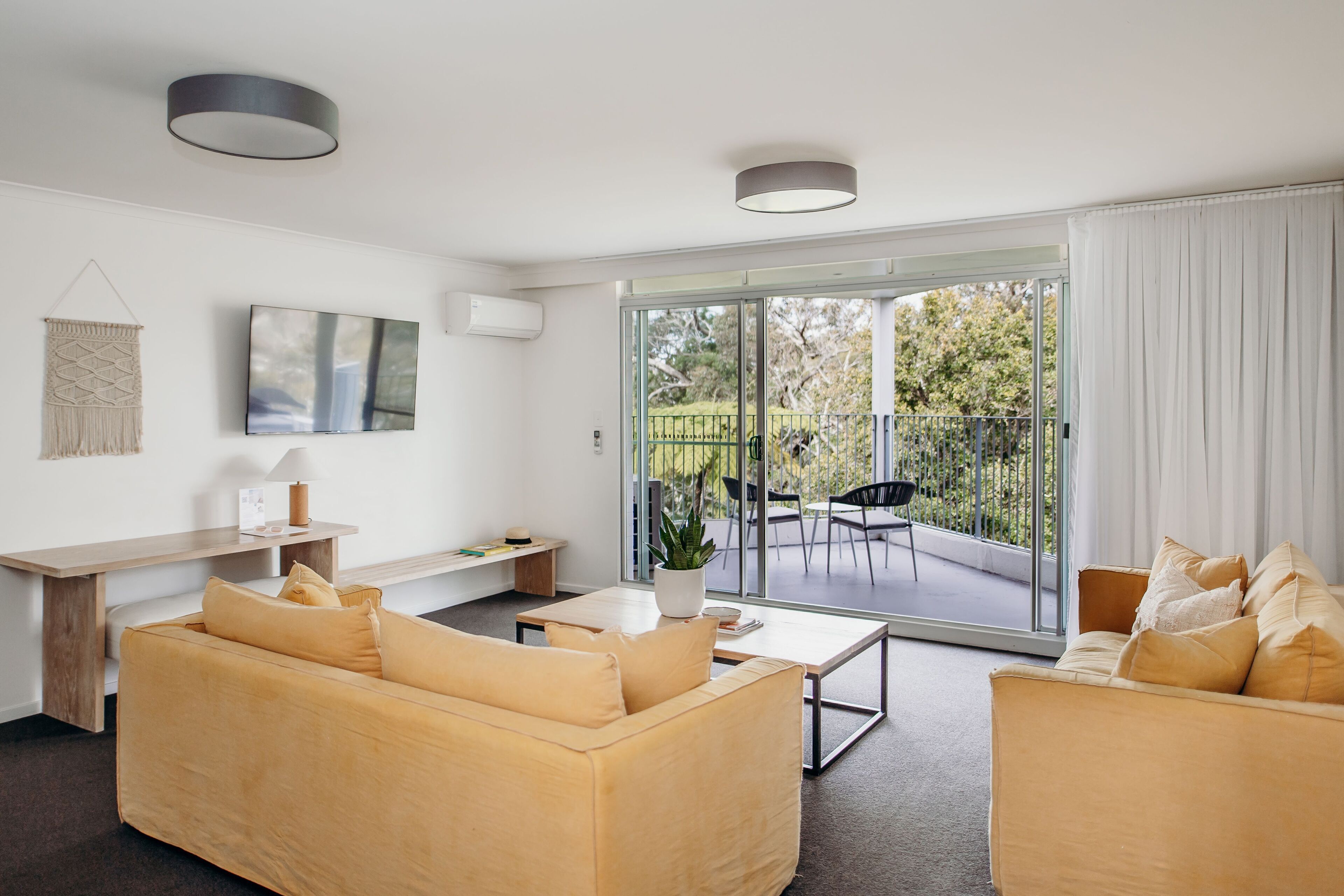 Una sala de estar moderna con un sofá, una mesa de centro y un televisor montado en la pared. Hay una puerta corrediza de vidrio que da a un patio exterior con una mesa y sillas.