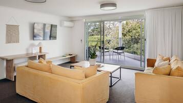 Una sala de estar moderna con un sofá, una mesa de centro y un televisor montado en la pared. Hay una puerta corrediza de vidrio que da a un patio exterior con una mesa y sillas.