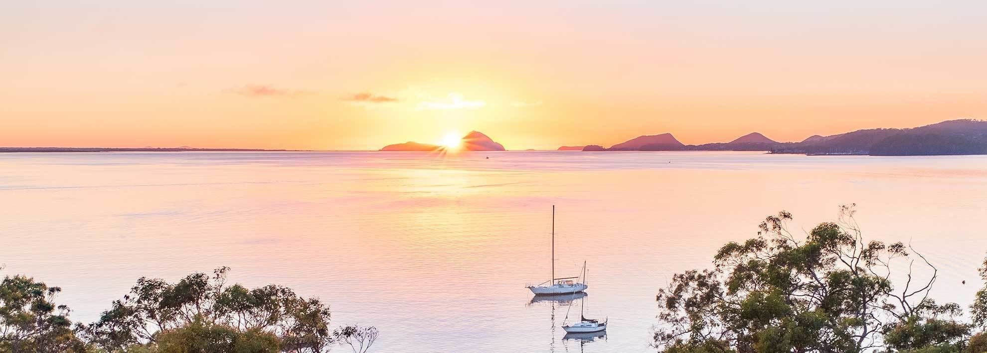 A calm body of water with boats, trees, and distant mountains during sunset.