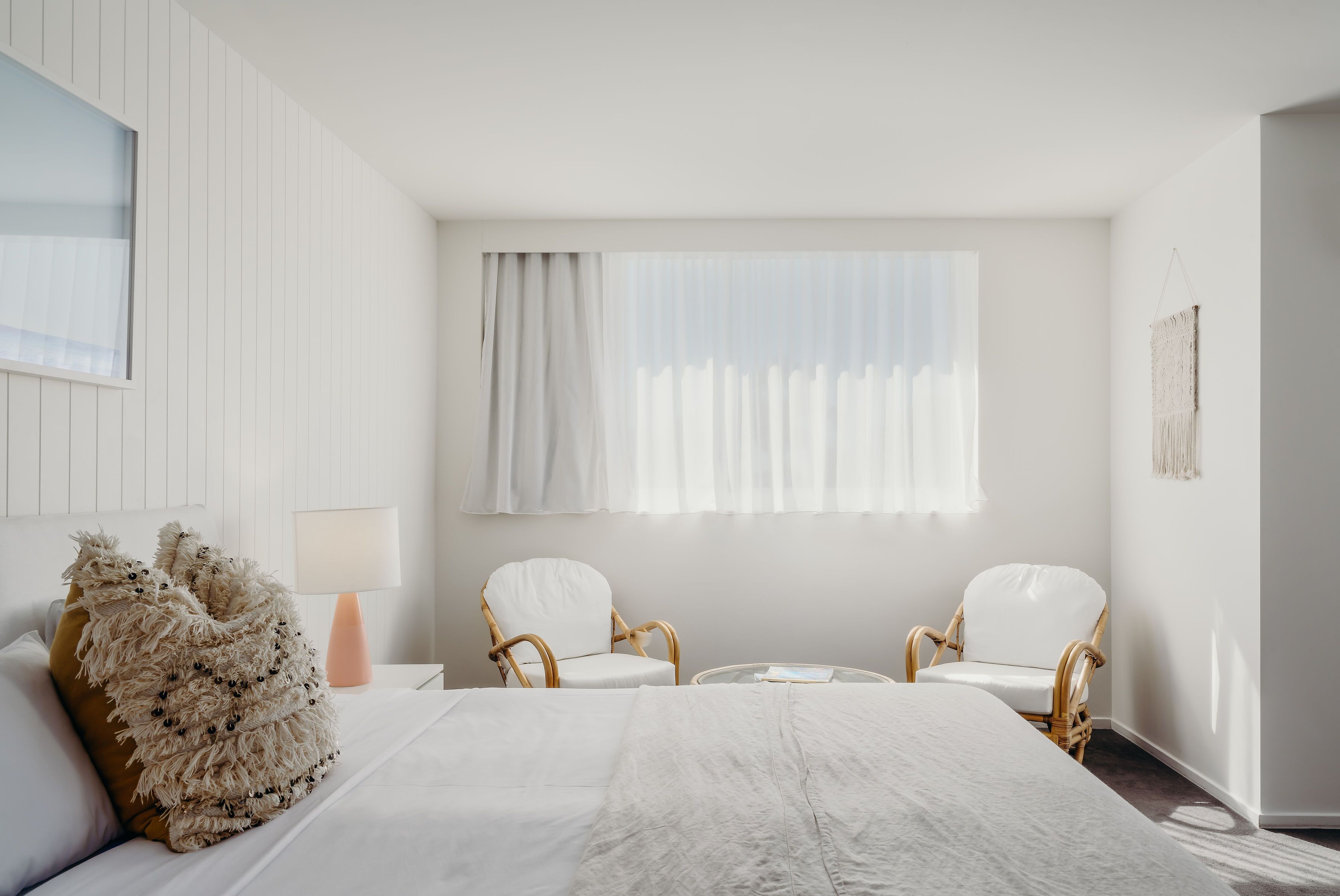 A hotel room with a large bed, two white armchairs, a small table, and a window with sheer curtains.