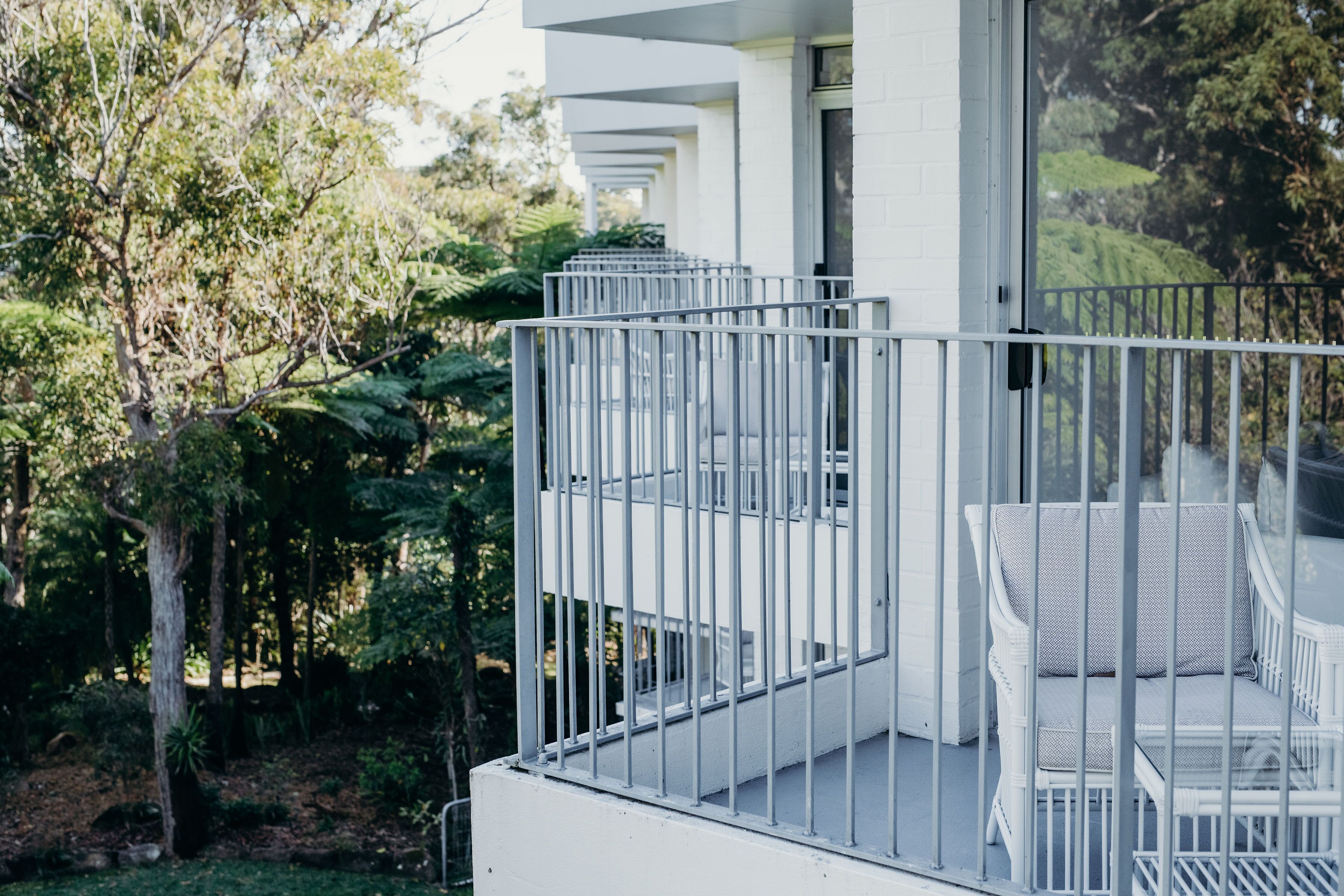 Uma varanda branca com grade de metal, uma cadeira de vime e vista para um jardim exuberante e árvores.