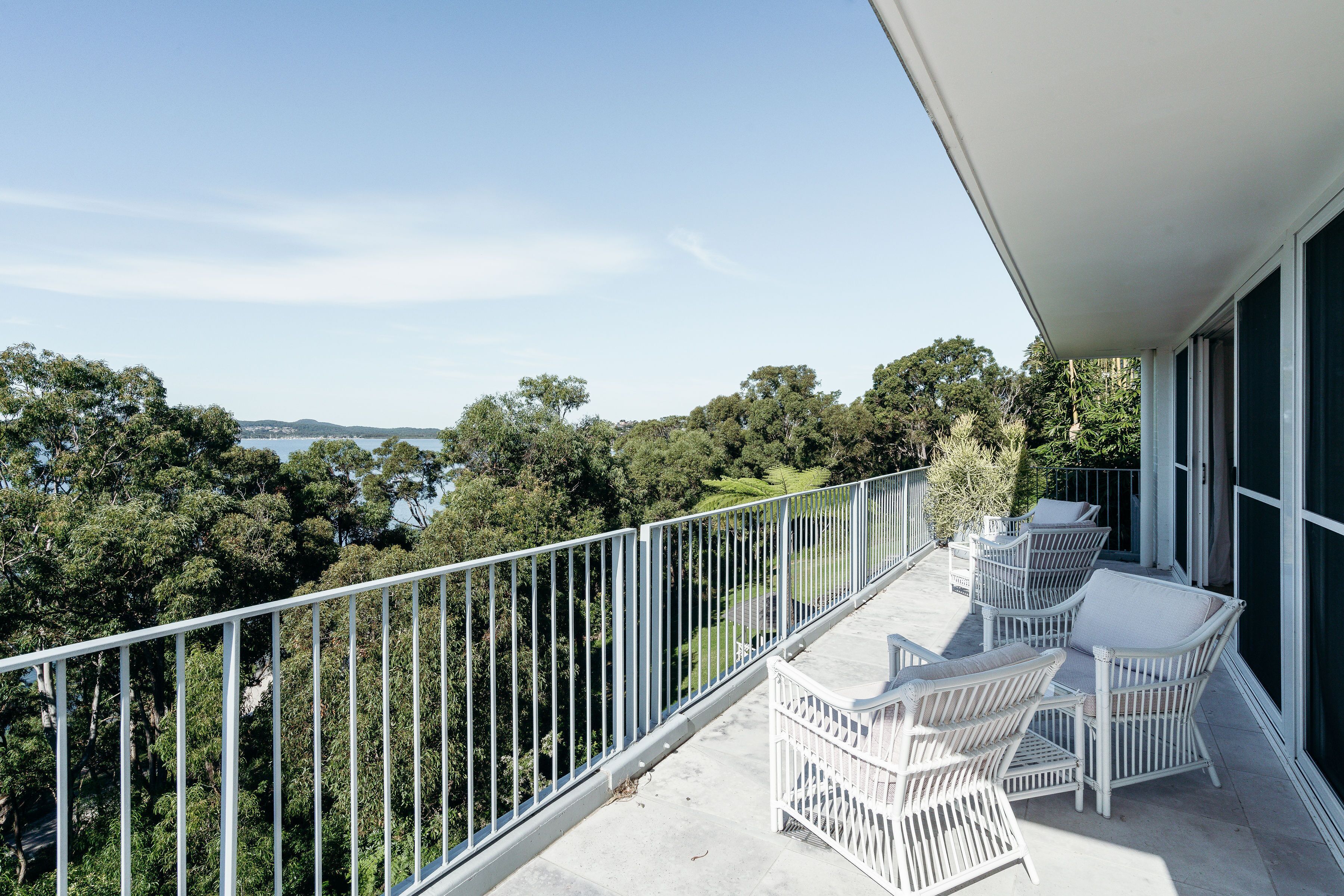A balcony with white wicker furniture and a view of a forested area and water.