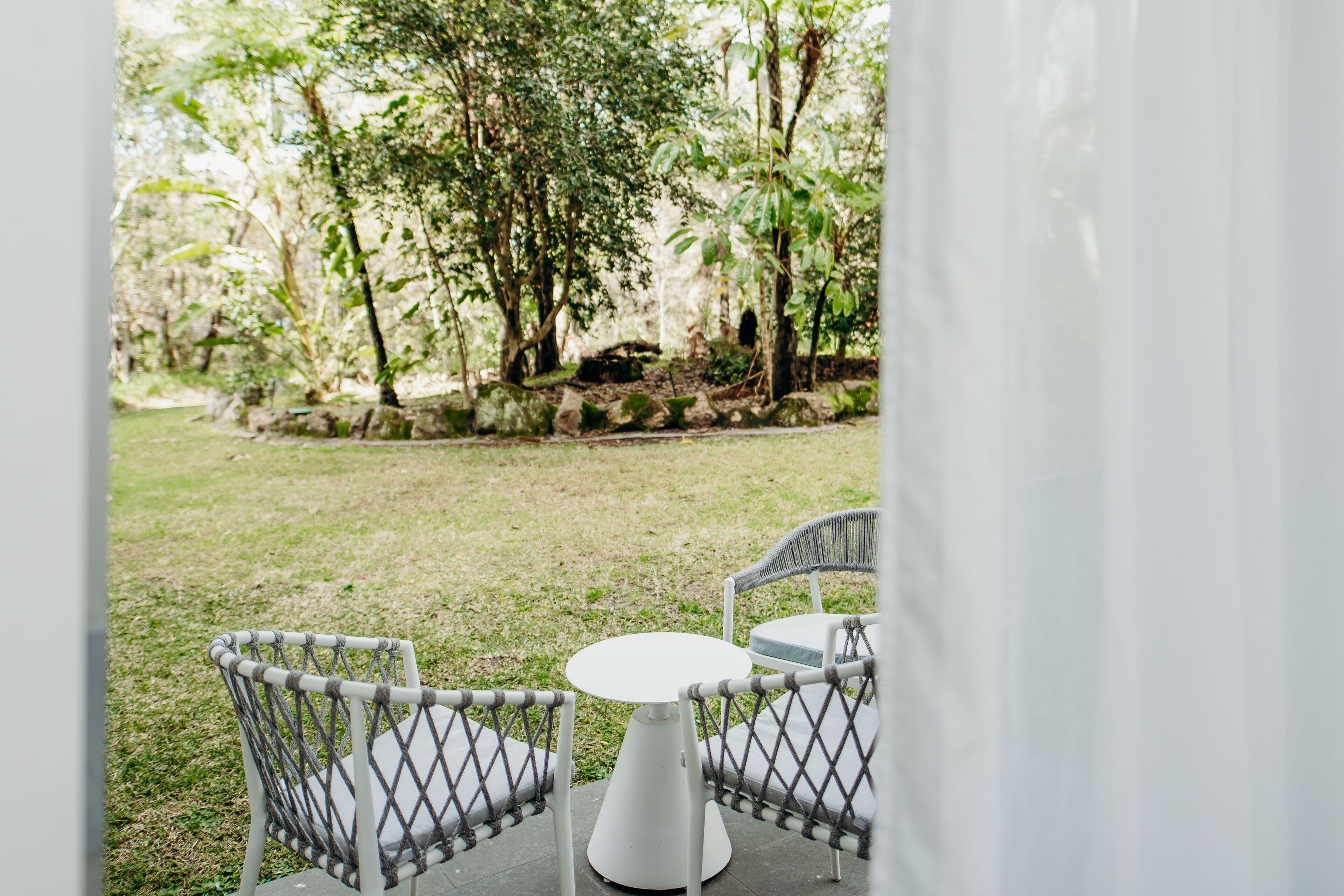 An outdoor seating area with a round table and two chairs, surrounded by a grassy lawn and trees.
