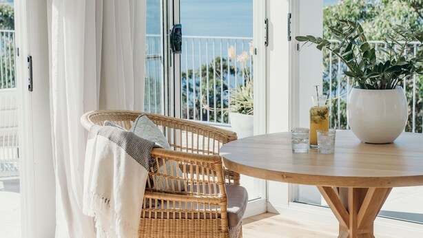 Un balcón con una silla de mimbre, una mesa redonda de madera y una planta en maceta.