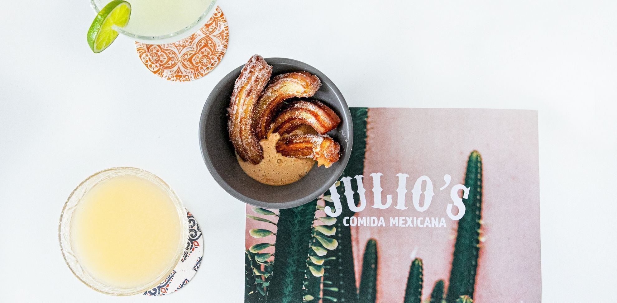 A table with churros, a glass of a yellow beverage, and a book titled "JULIO'S COMIDA MEXICANA".