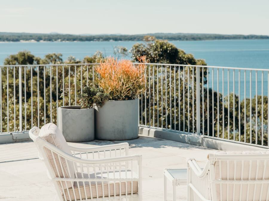 A balcony with white wicker chairs and a potted plant, overlooking a body of water and trees.