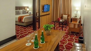 Habitación de hotel con cama, televisor, mesa de madera con vasos y una planta, y alfombra roja con estampado.