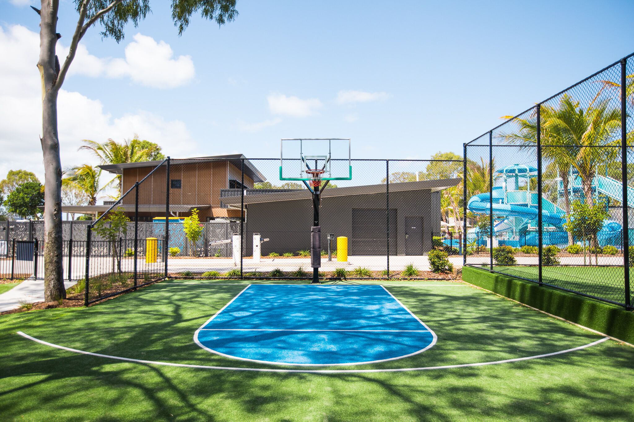 An outdoor basketball court with a blue surface, surrounded by a fence, trees, and a building in the background.