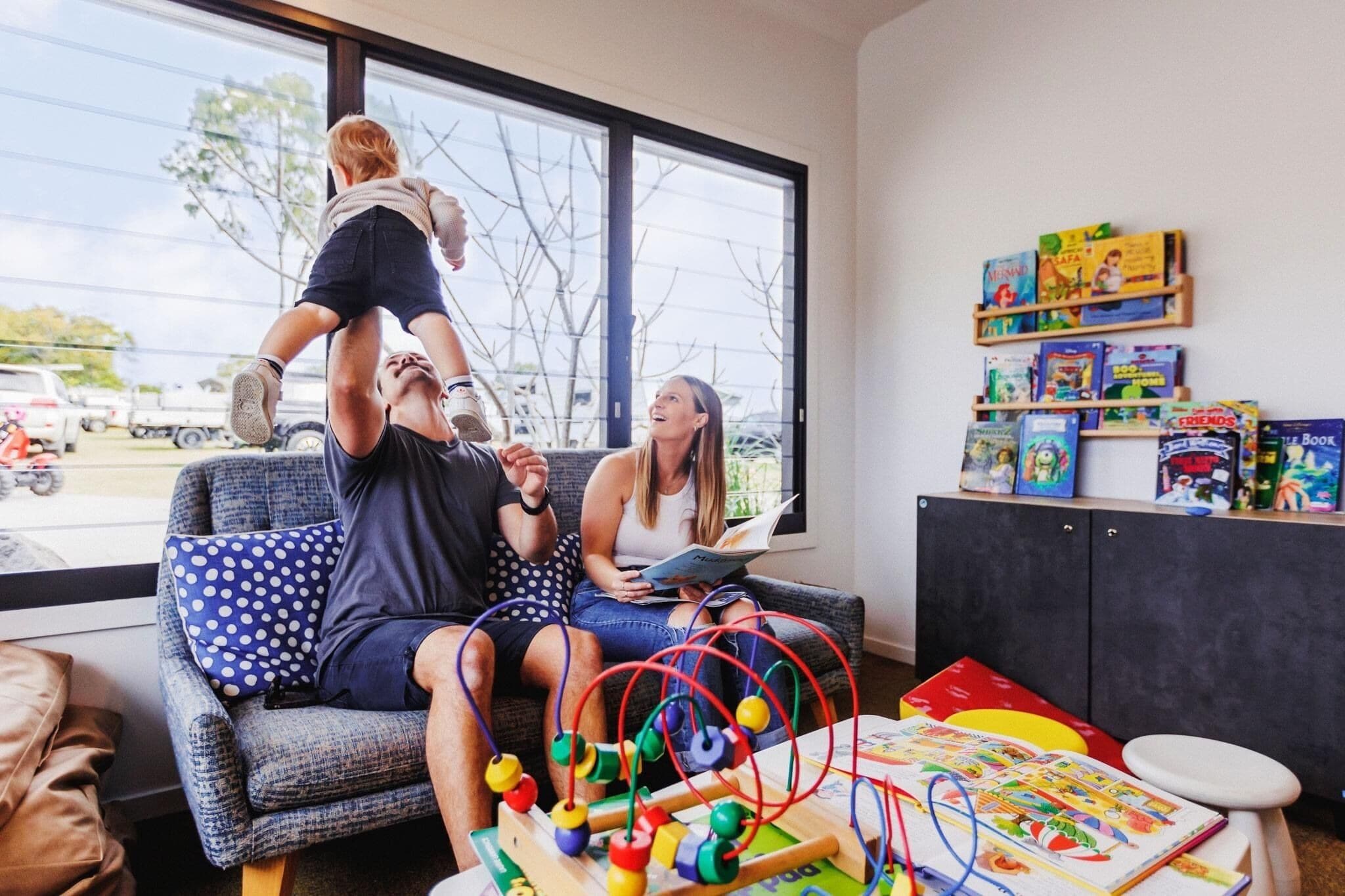 Een kamer met een groot raam, een bank, een baby op de schouders van de man, en een boekenkast met kinderboeken.