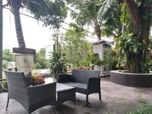 Un espacio de estar al aire libre cubierto con mobiliario de mimbre, rodeado de abundante vegetación y una palmera.