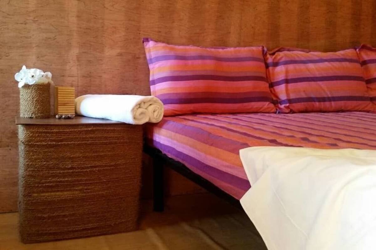 Un lit avec une literie à rayures, une table de chevet en bois sur laquelle est posée une serviette enroulée, et un petit objet décoratif.