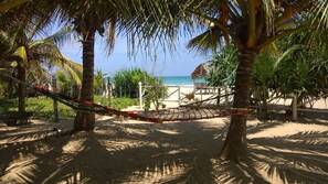A hammock between palm trees overlooking a beach with a thatched-roof structure in the background.