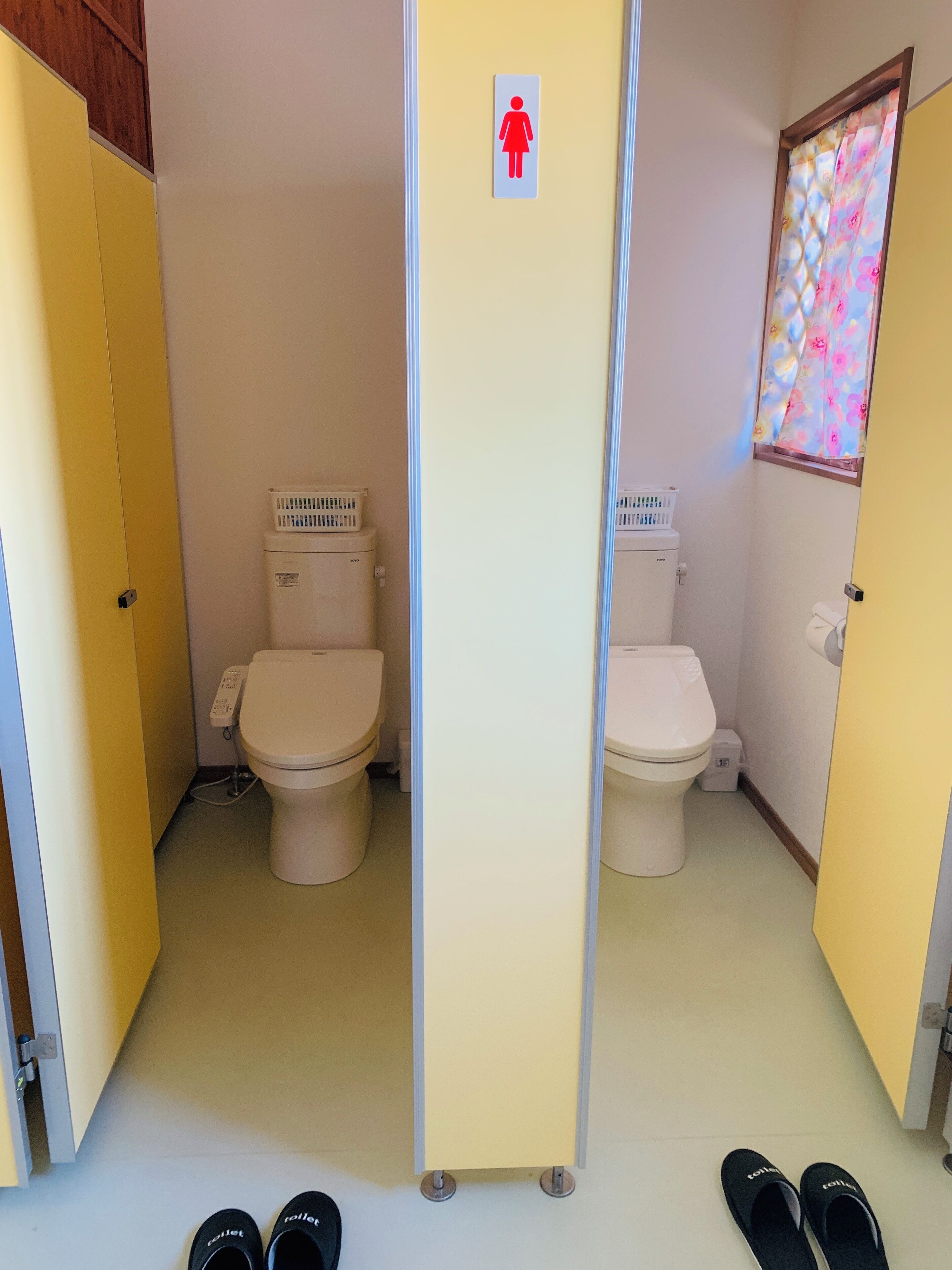 Baño con dos inodoros, separador para mayor privacidad y ventana con cortina floral.