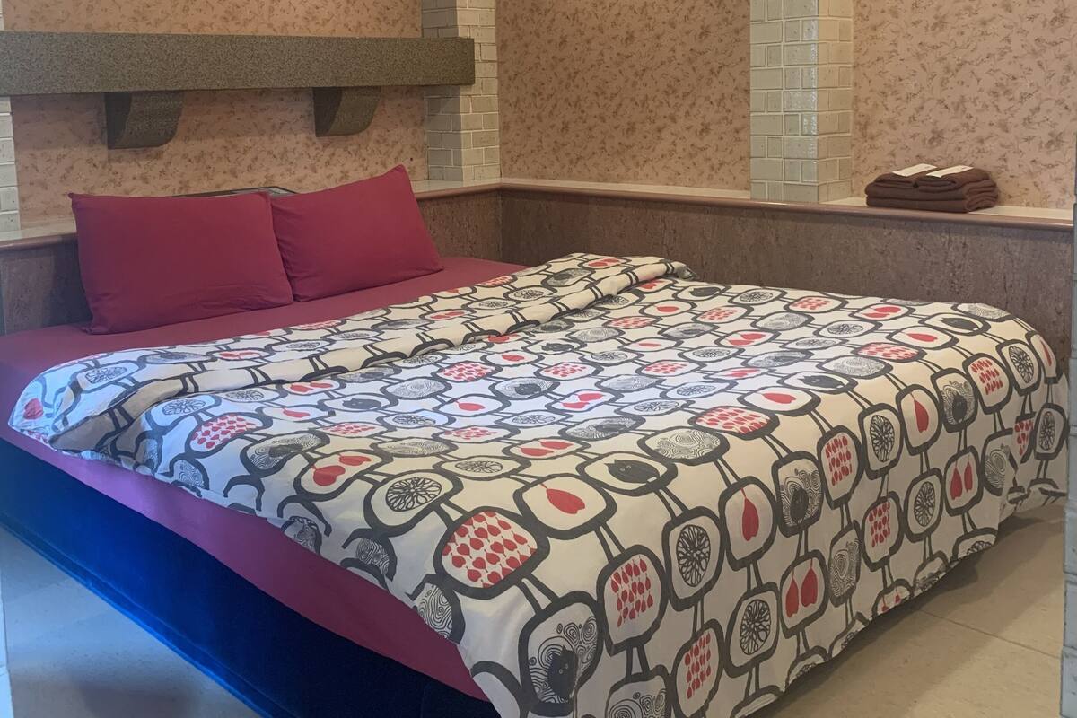 A bedroom with a patterned bedspread, red pillows, and a brick-patterned wall.