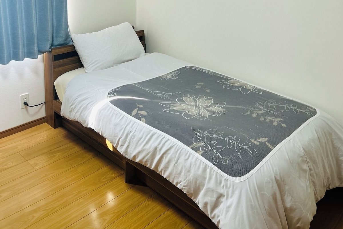 A single bed with a floral-patterned bedspread, a wooden floor, a blue curtain, and a wall-mounted air conditioner.
