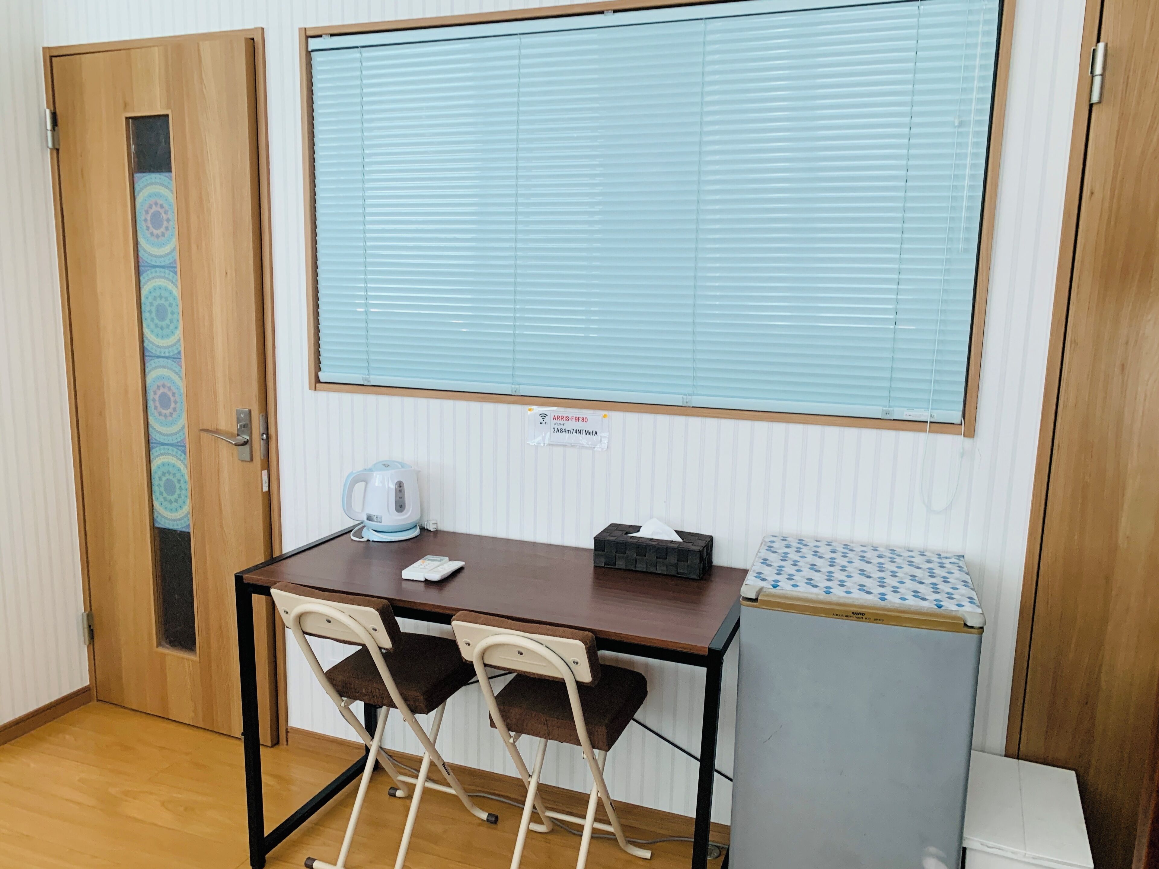 A small room with a wooden desk, two chairs, a kettle, a tissue box, a small refrigerator, and a window with blinds.