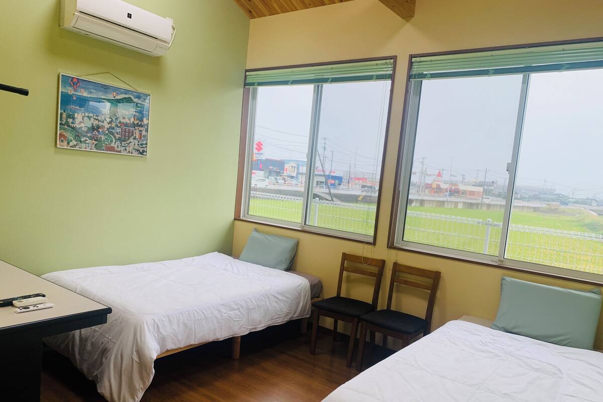 A room with two beds, a desk, and a chair. There are two windows with blinds, and a painting on the wall.