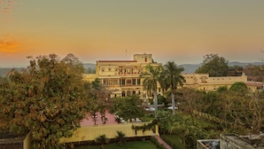 A large, historic building with multiple balconies and a flag, surrounded by lush greenery and a clear sky. - Fort Dhariyawad Heritage Hotel Udaipur (Dhariyawad)