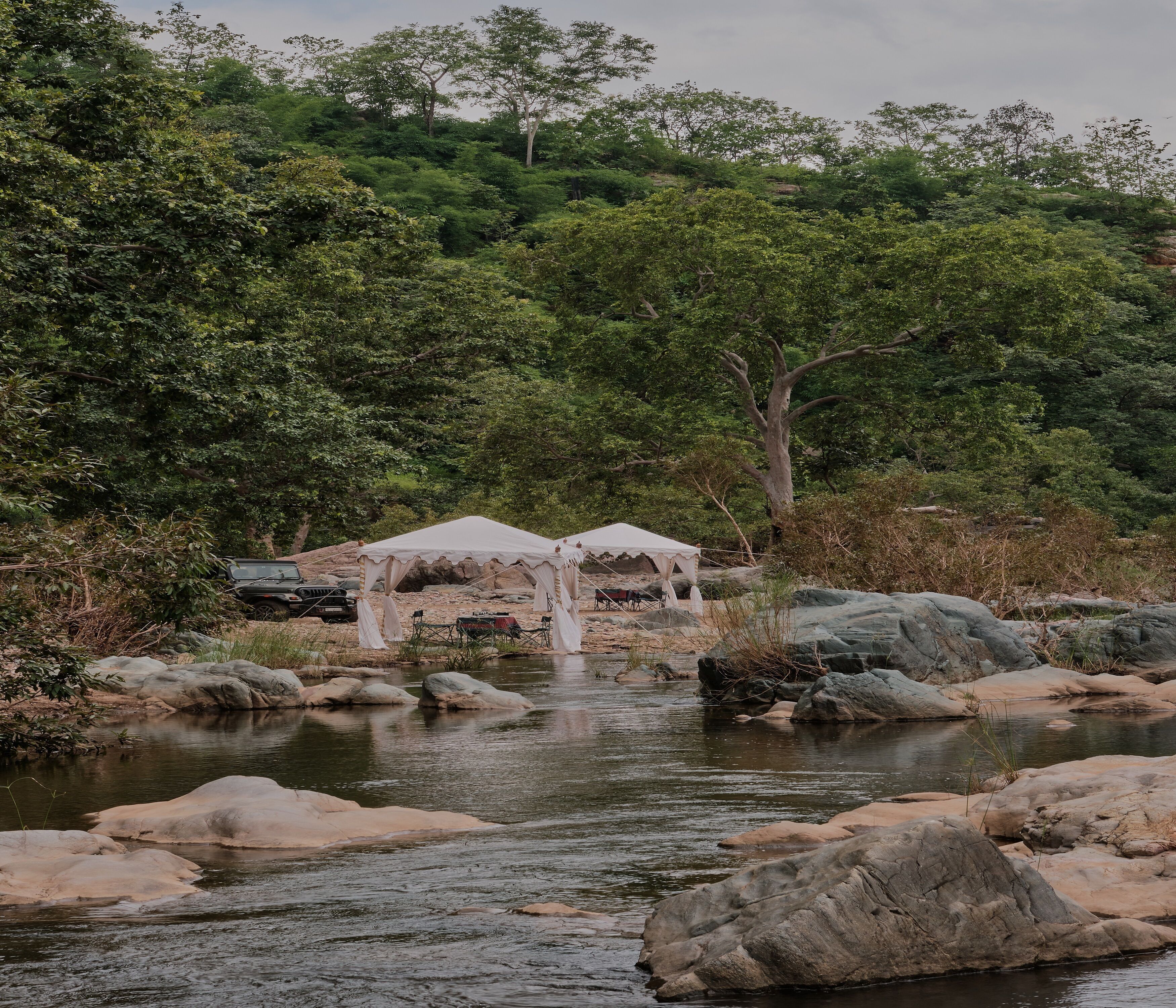 A river with rocks, two white tents, and a forested hillside.