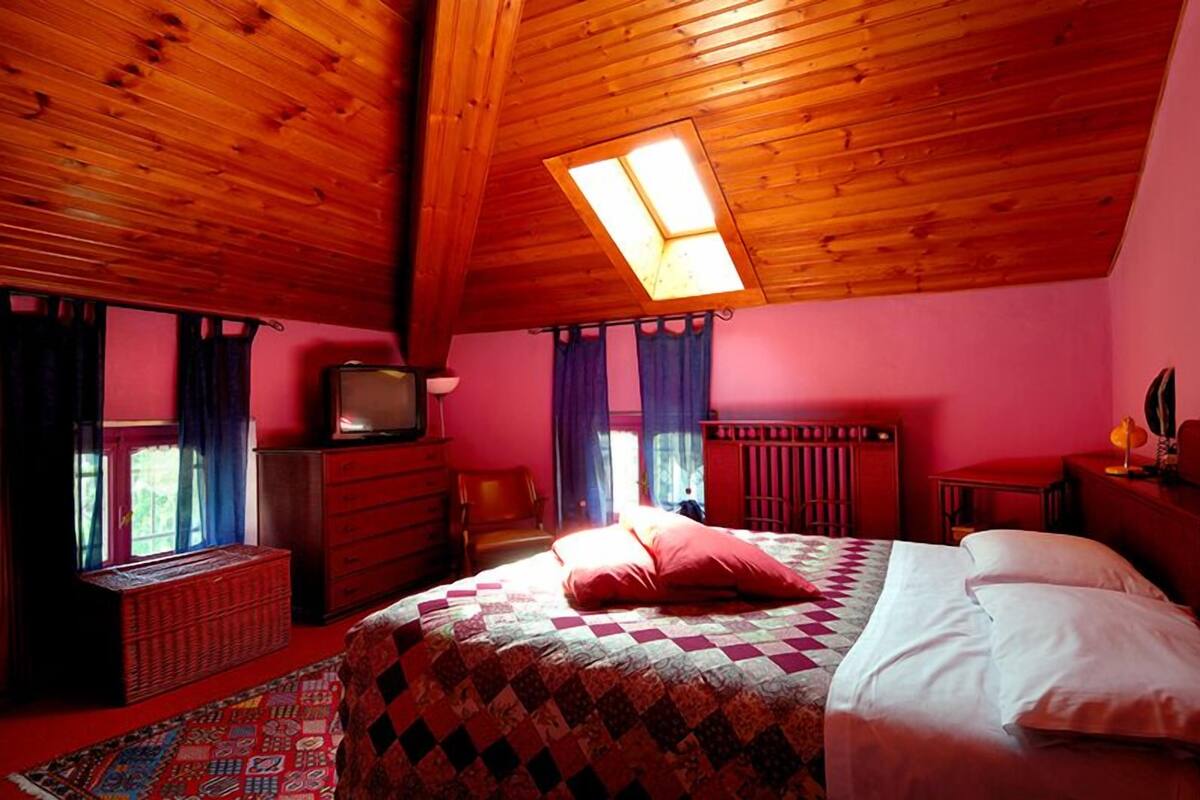 A bedroom with a wooden ceiling, a bed with a checkered quilt, a television on a wooden dresser, and a window with curtains.