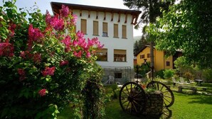 Un edificio con fachada blanca y varias ventanas, rodeado de abundante vegetación y flores rosadas.