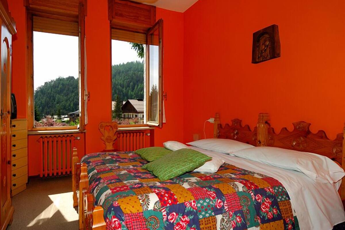 A bedroom with a wooden bed, a colorful quilt, a window with a view of a forest, and a red wall.