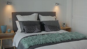 Una cama bien hecha con un cabecero gris, varios almohadones, un florero azul y una mesita de noche con un marco de fotos.