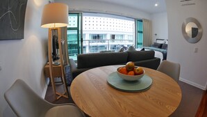 Una habitación de hotel moderna con una mesa redonda para comer, un sofá, una lámpara de pie y un amplio ventanal con vistas a un edificio.