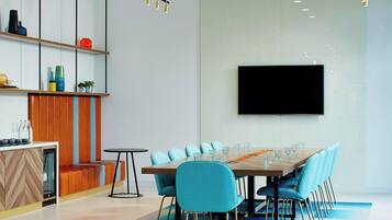 A modern conference room with a long table, blue chairs, a TV screen, and a wooden partition with shelves.