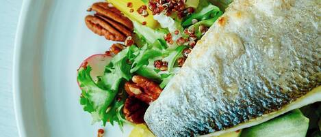 Un plato blanco con un pescado a la parrilla, rodajas de aguacate, rábanos y nueces sobre una mesa de madera.