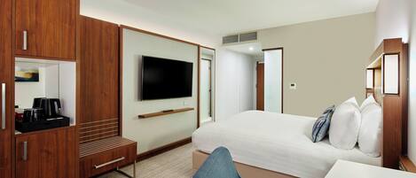 A hotel room with a large bed, a TV mounted on the wall, a wooden wardrobe, a desk with a chair, and a small seating area.