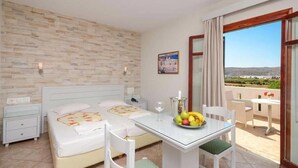A hotel room with two beds, a dining table with fruit, a balcony with a view, and a brick-patterned wall. - Plaka Hotel I (Naxos)