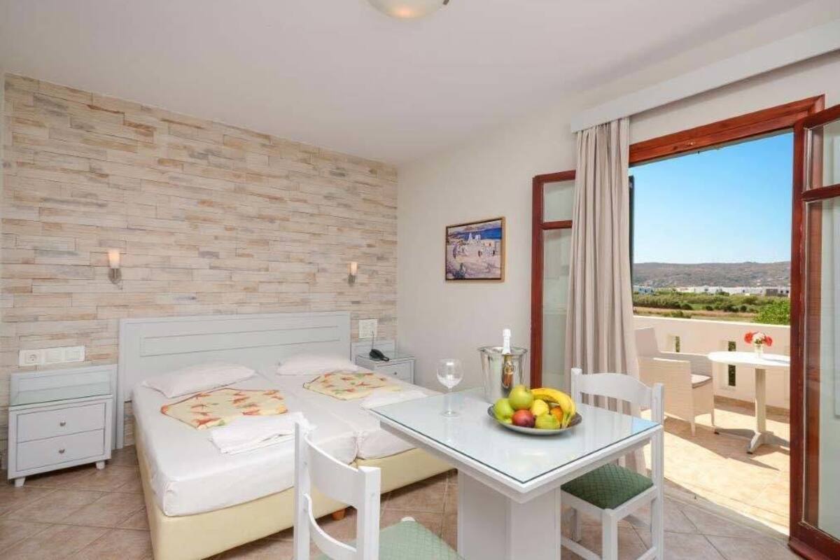A hotel room with two beds, a dining table with fruit, a balcony with a view, and a brick-patterned wall.