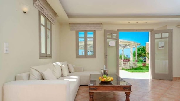 A living room with a sofa, a glass coffee table with fruit, and a view of a garden through open doors. - Plaka Hotel I (Naxos)