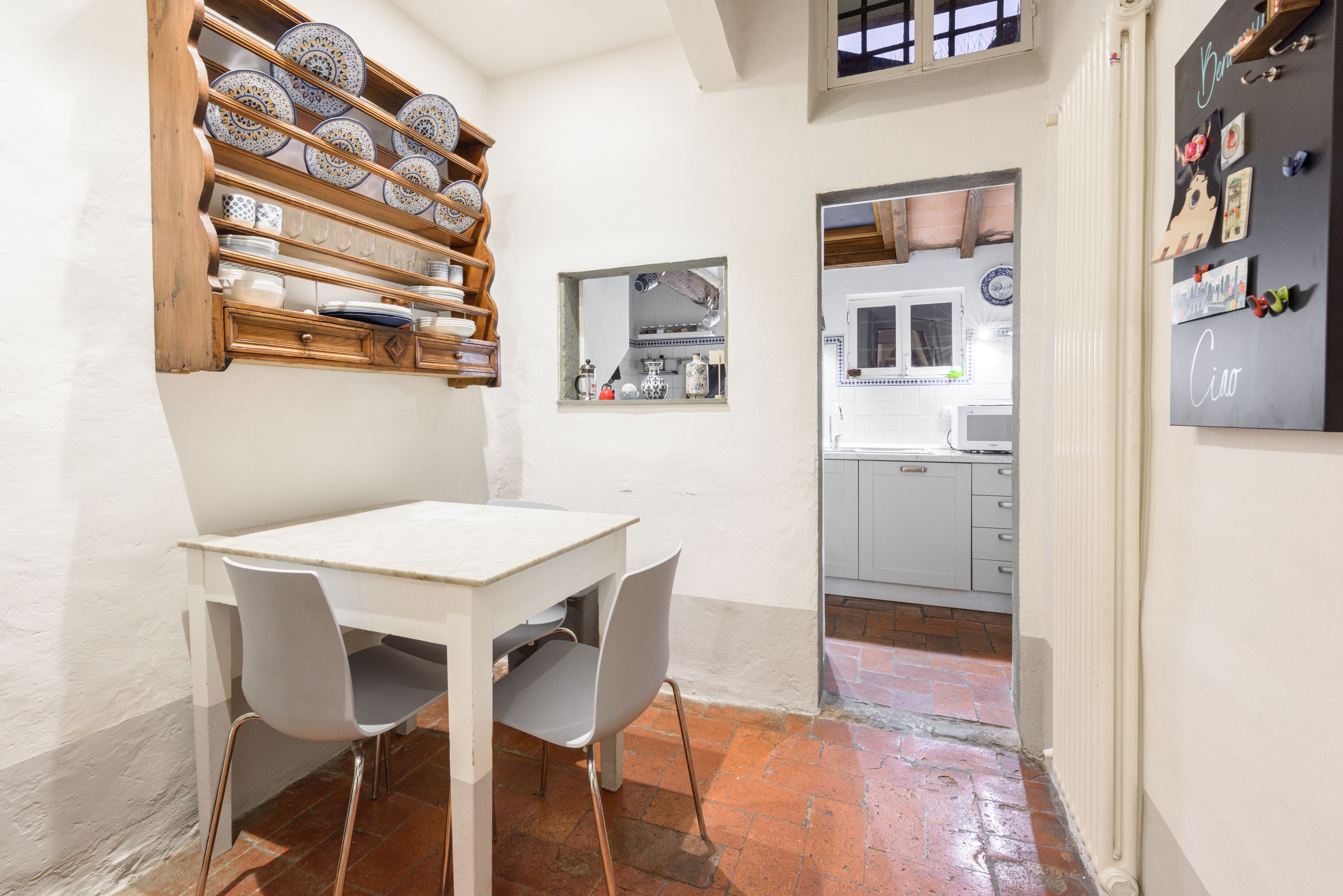 A dining area with a white table and chairs, a wooden shelf with plates, a mirror, and a kitchen in the background.