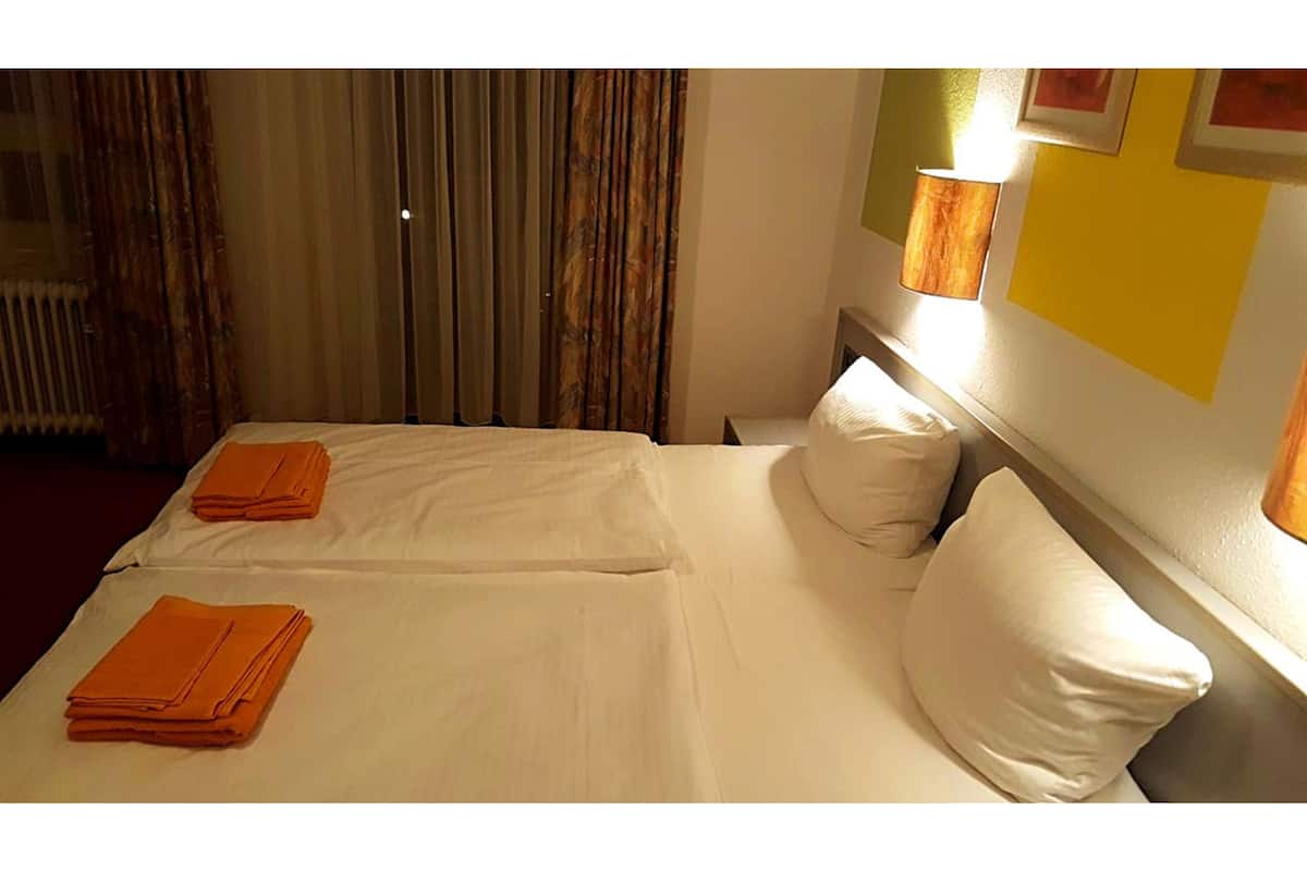 A hotel room with two beds, a radiator, curtains, and a wall-mounted lamp.