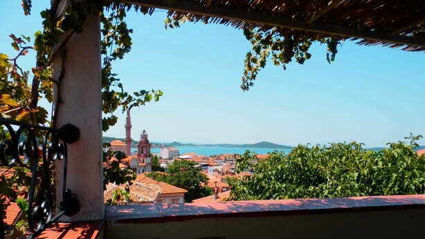 A balcony with a view of a coastal town, including a church tower and buildings with red-tiled roofs. - Taksiyarhis Pension (Ayvalik)