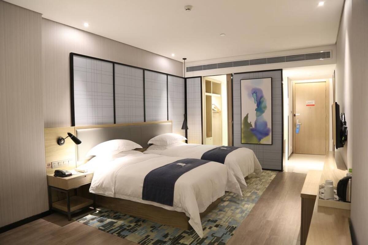 A hotel room with two beds, a bedside table with a lamp, a large artwork on the wall, and a view of the hallway.