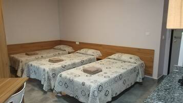 A small, clean room with three single beds, a wooden headboard, a small table, and a kitchenette area.