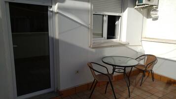A small patio area with a glass table and two wicker chairs, a sliding glass door, and a window with a white blind.