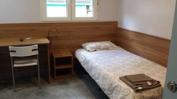 A small, single-room accommodation with a wooden desk, chair, and a bed with a patterned duvet.