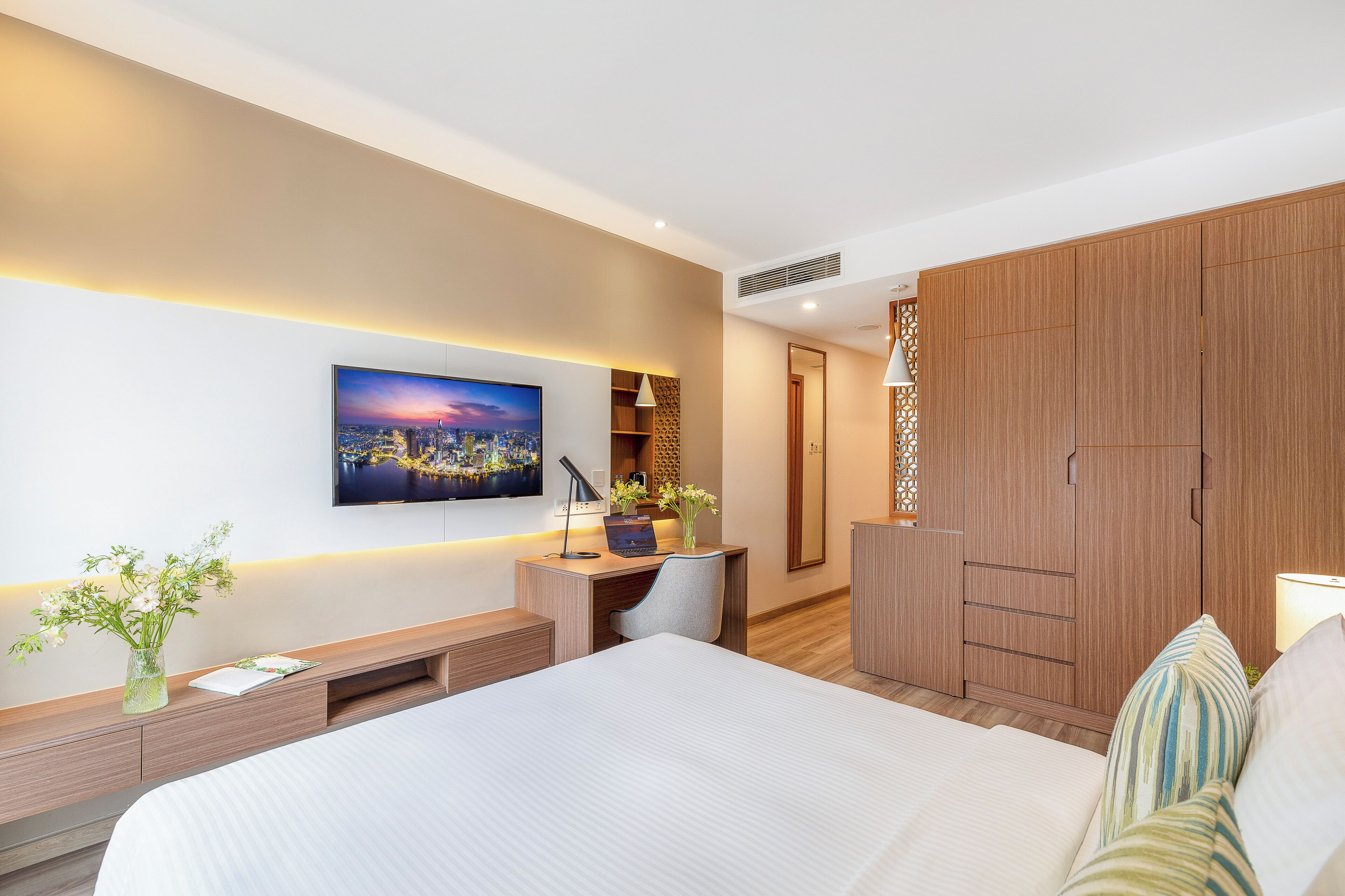 Una habitación de hotel moderna con una cama grande, un escritorio con una computadora portátil, un televisor montado en la pared y un armario de madera.